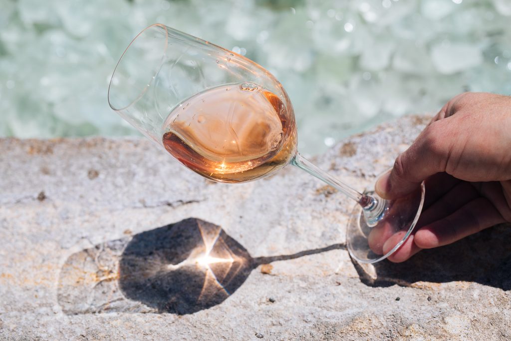 male hand holding a glass of rosé wine at an angle in front of a water feature
